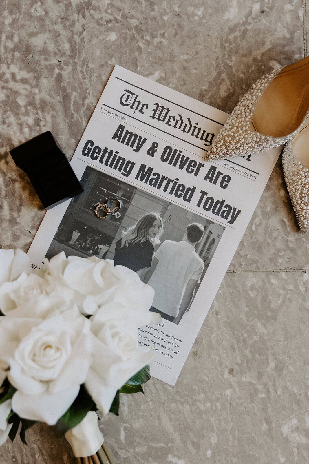Elegant wedding shoe with pearl embellishments on a marble floor, surrounded by wedding rings, a bouquet of white roses, and a newspaper wedding announcement.