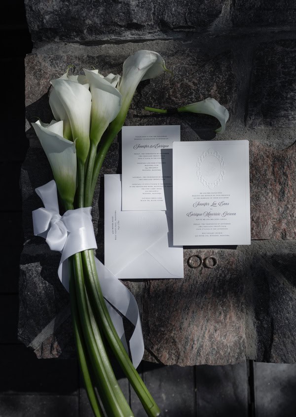 White calla lilies bouquet with wedding invitation and rings on a stone surface.