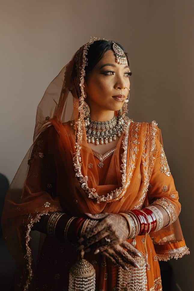 Elegant Indian bride wearing traditional orange bridal outfit with intricate jewelry and henna designs on her hands.