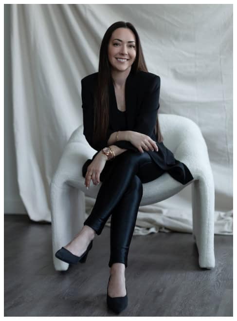 Melanie Parente sitting on a white modern chair, dressed in a black blazer and leather pants, smiling confidently, at her Winnipeg wedding and event planning studio.