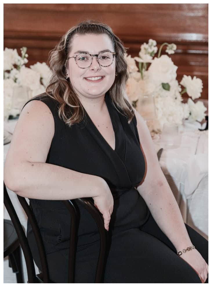 Elegant woman in black dress sitting at wedding or event reception with floral centerpieces, smiling, in a rustic wooden venue.