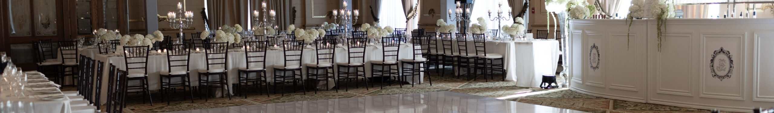 Elegant wedding reception setup with round tables, white linens, floral centerpieces, and black chairs in a sophisticated banquet hall.