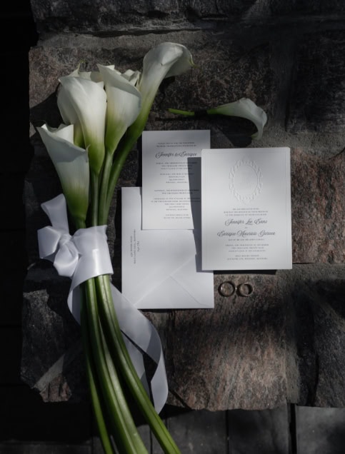 White calla lilies bridal bouquet with wedding invitations and rings on a stone surface.