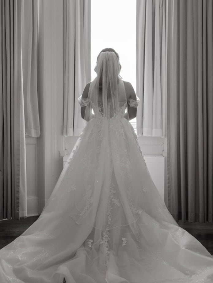 Elegant bride standing in front of a window with curtains, wearing a beautiful wedding gown with lace details and a veil, ready for her wedding day.